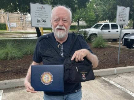 Man posed outdoors holding a certificate holder and lunch bag