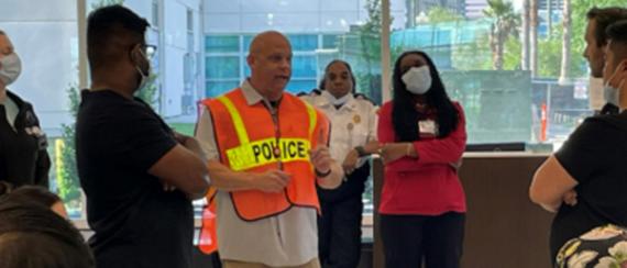 Candid image of a group of people gathered around an officer who is speaking to everyone and wearing a bright orange 'Police' safety vest.