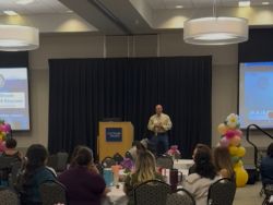 Man standing on stage speaking to crowd in a room