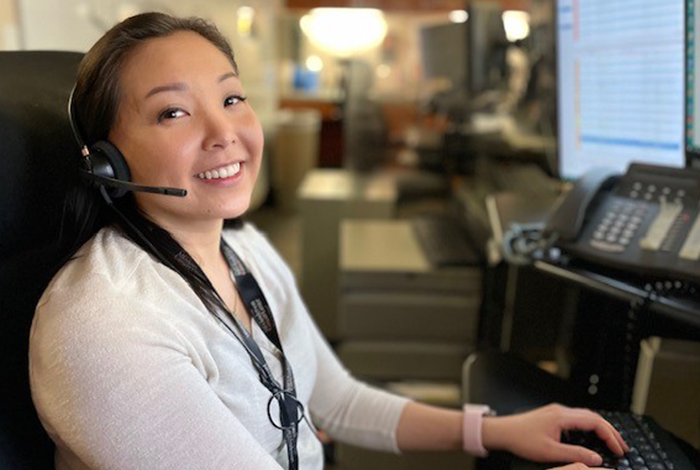 Woman wearing long sleeved beige shirt smiling at the camera with her teeth showing with a headset on receiving calls from the dispatch department with many computer screens in front of her. Fingers are on the keyboard in ready position to type.