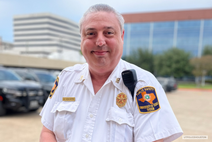 Senior Public Safety Officer Armando Viscarri wearing his UT Police uniform white button down shirt with police patch, badge, and radio on his left shoulder. he is smiling into the camera with a blurred background